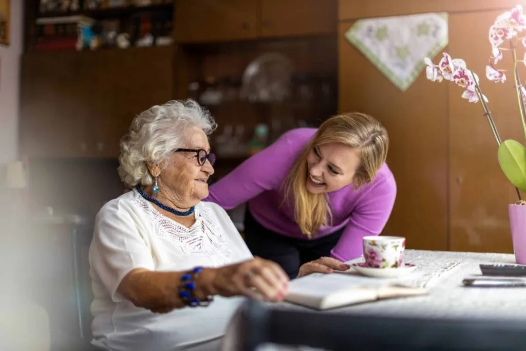 Smiling female carer supporting an older woman at the kitchen table, sharing a cup of tea and conversation to promote dignity and wellbeing in care.